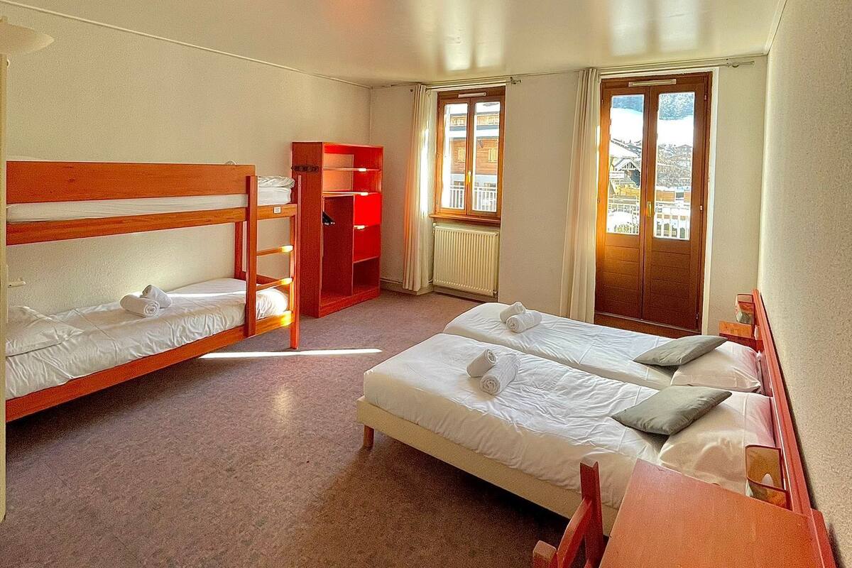 Une chambre avec deux lits superposés, un bureau en bois, une armoire rouge et une fenêtre donnant sur des immeubles.