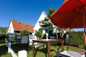 Un espace extérieur aménagé avec des tables et des chaises, un parasol rouge et, en arrière-plan, un bâtiment blanc avec un toit rouge.