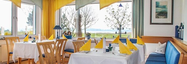 Un espace repas avec des tables préparées pour deux personnes, des chaises et une vue sur l’extérieur.