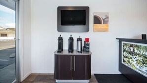 Un área de recepción con una televisión de pantalla plana montada en la pared, una estación de café con varios dispensadores y una pantalla digital mostrando un mensaje.