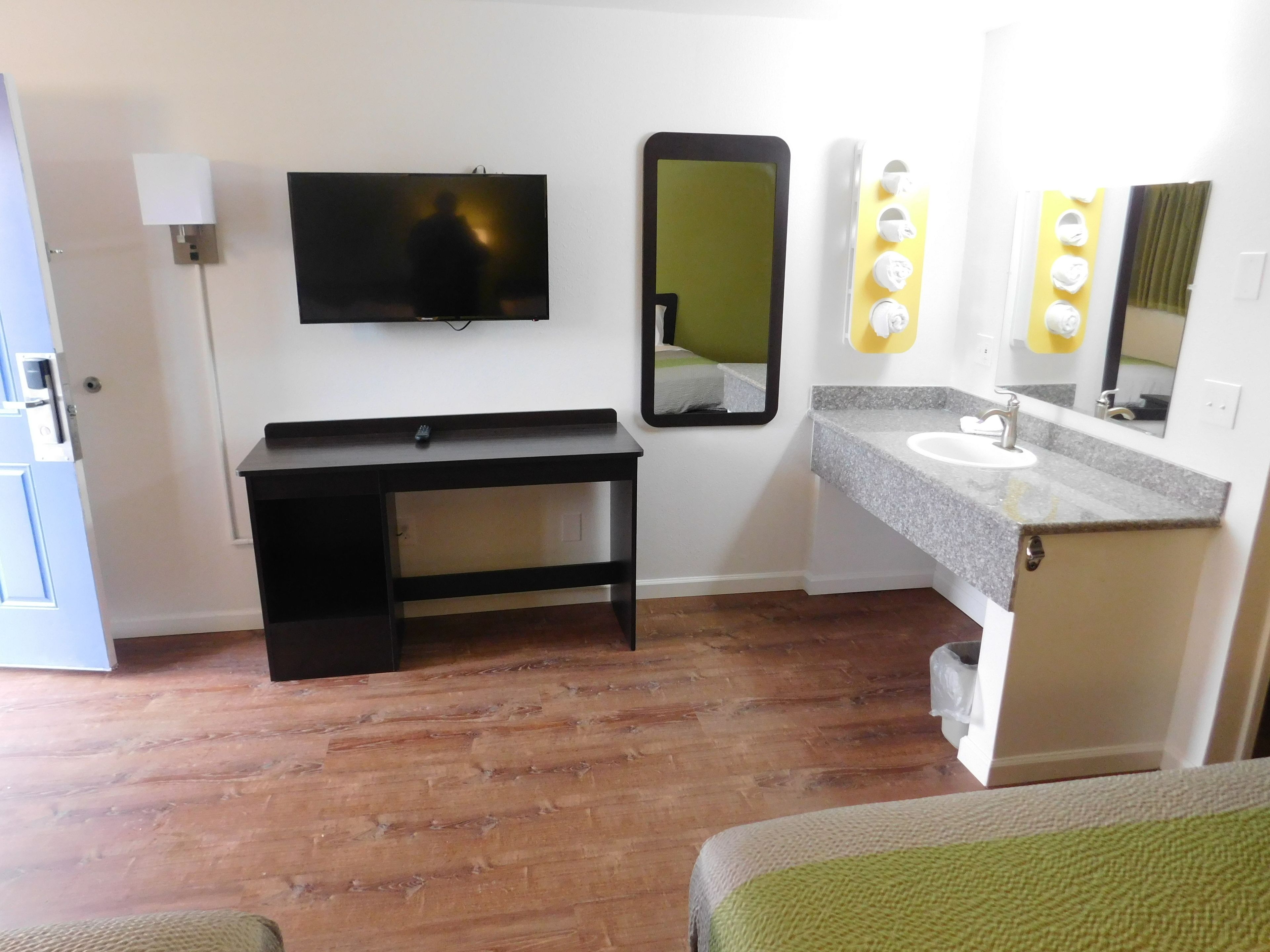 A hotel room with a wooden floor, a desk with a flat-screen TV, a large mirror, a bathroom with a sink and vanity, and a bed with a green blanket.