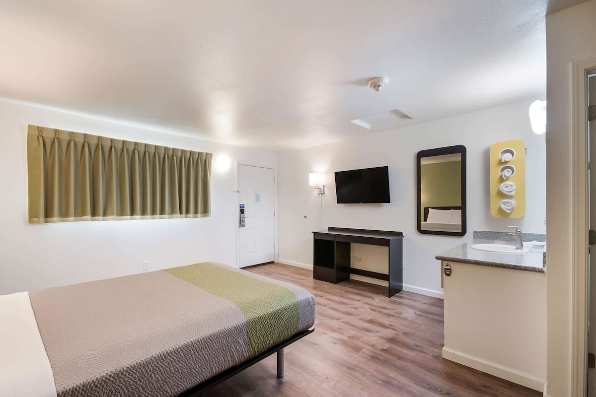 A hotel room with a bed, a desk, a TV, a mirror, and a bathroom with a sink and towel rack.