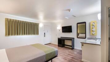 A hotel room with a bed, a desk, a TV, a mirror, and a bathroom with a sink and towel rack.