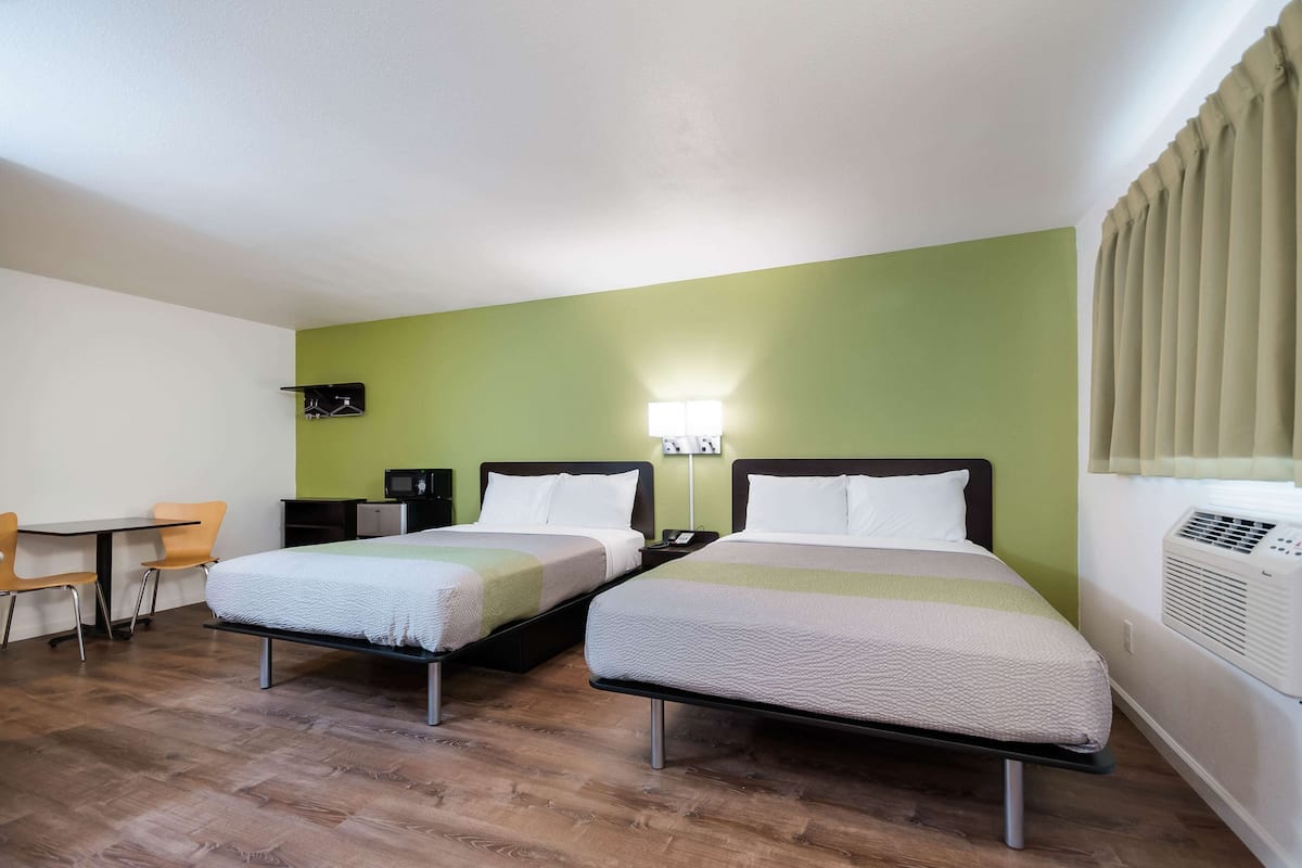A hotel room with two beds, a desk, chairs, a microwave, and an air conditioning unit.