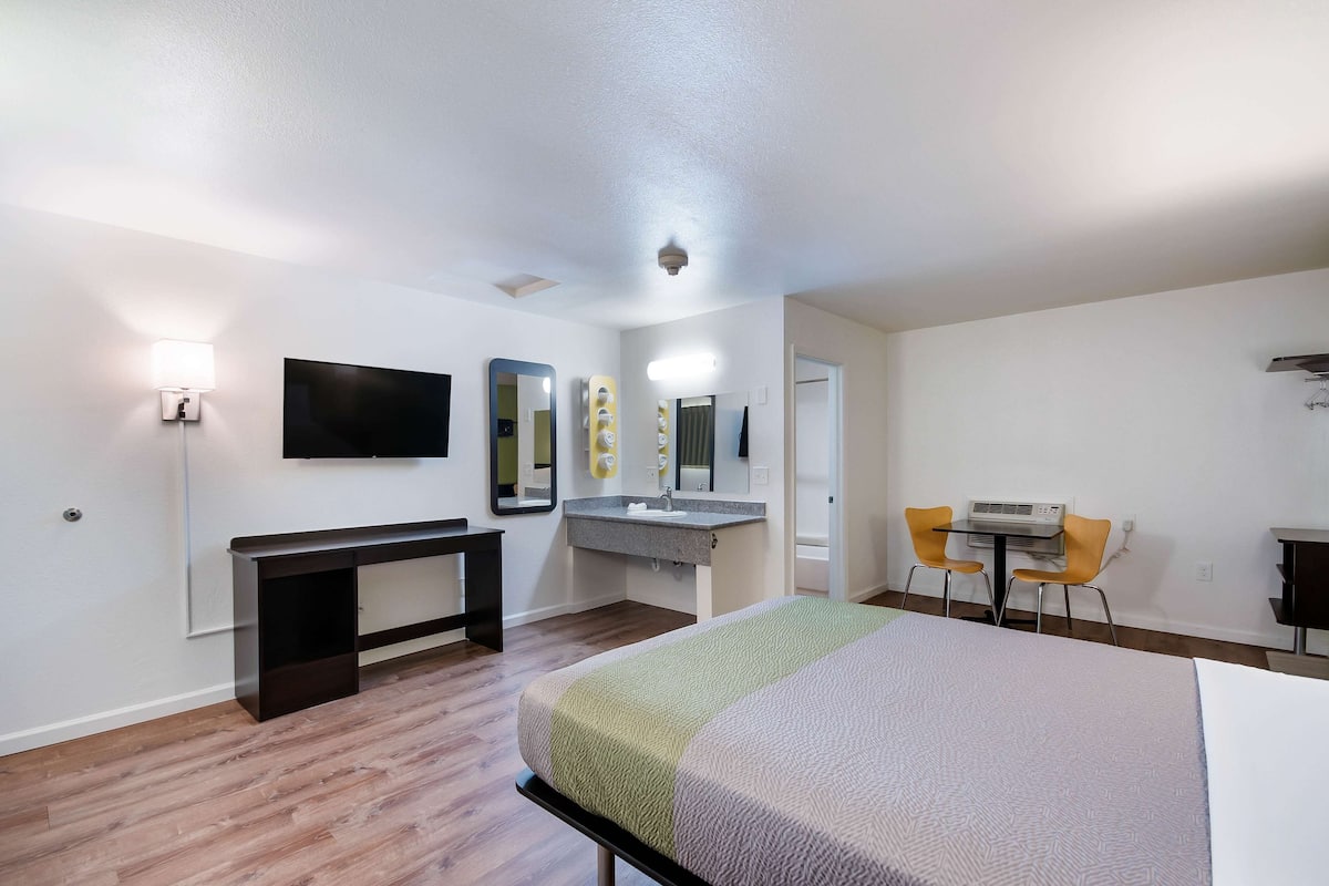 A hotel room with a bed, a desk, a chair, a TV, a mirror, and a bathroom.