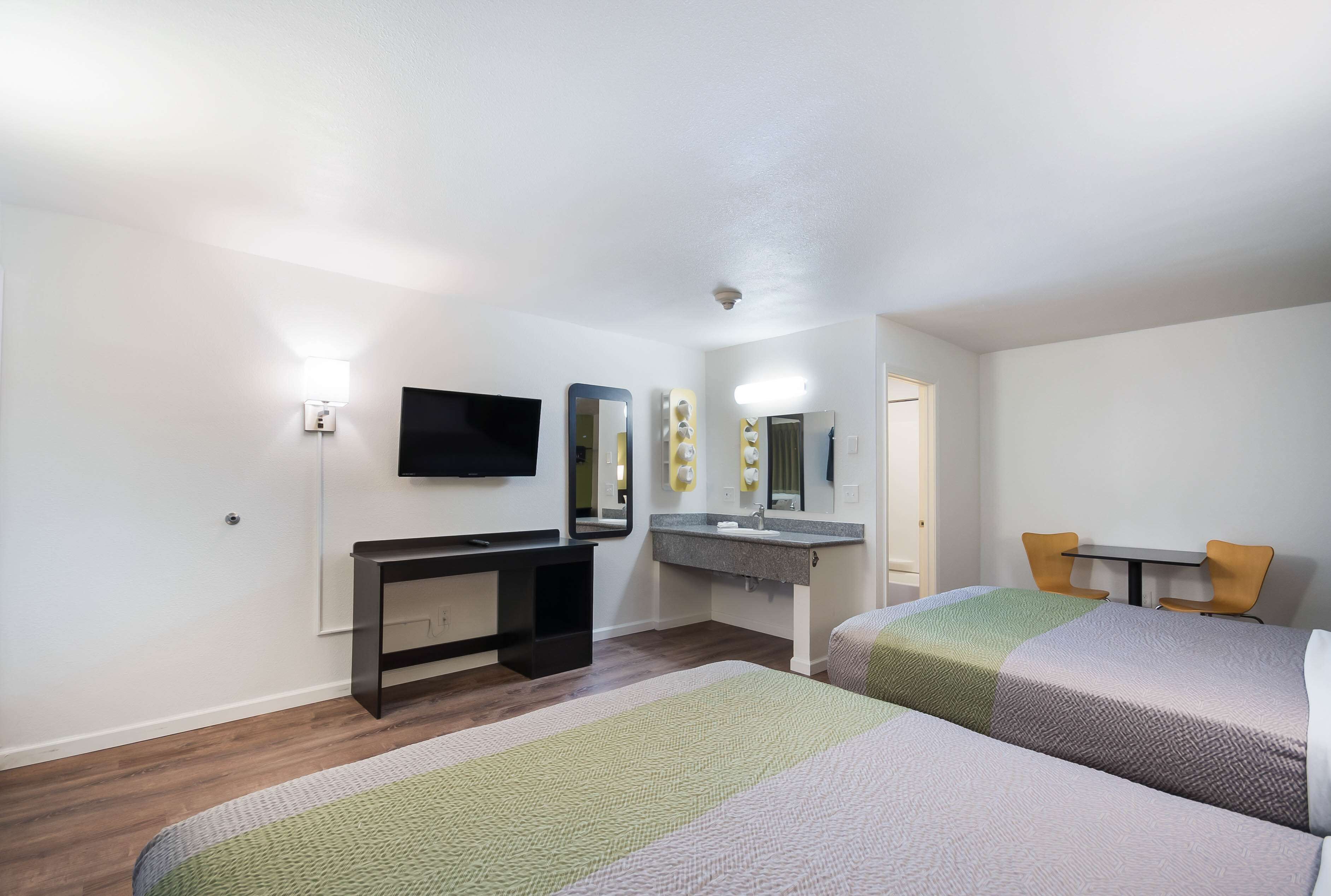 A hotel room with two beds, a desk, a TV, a bathroom mirror, and a small table with chairs.