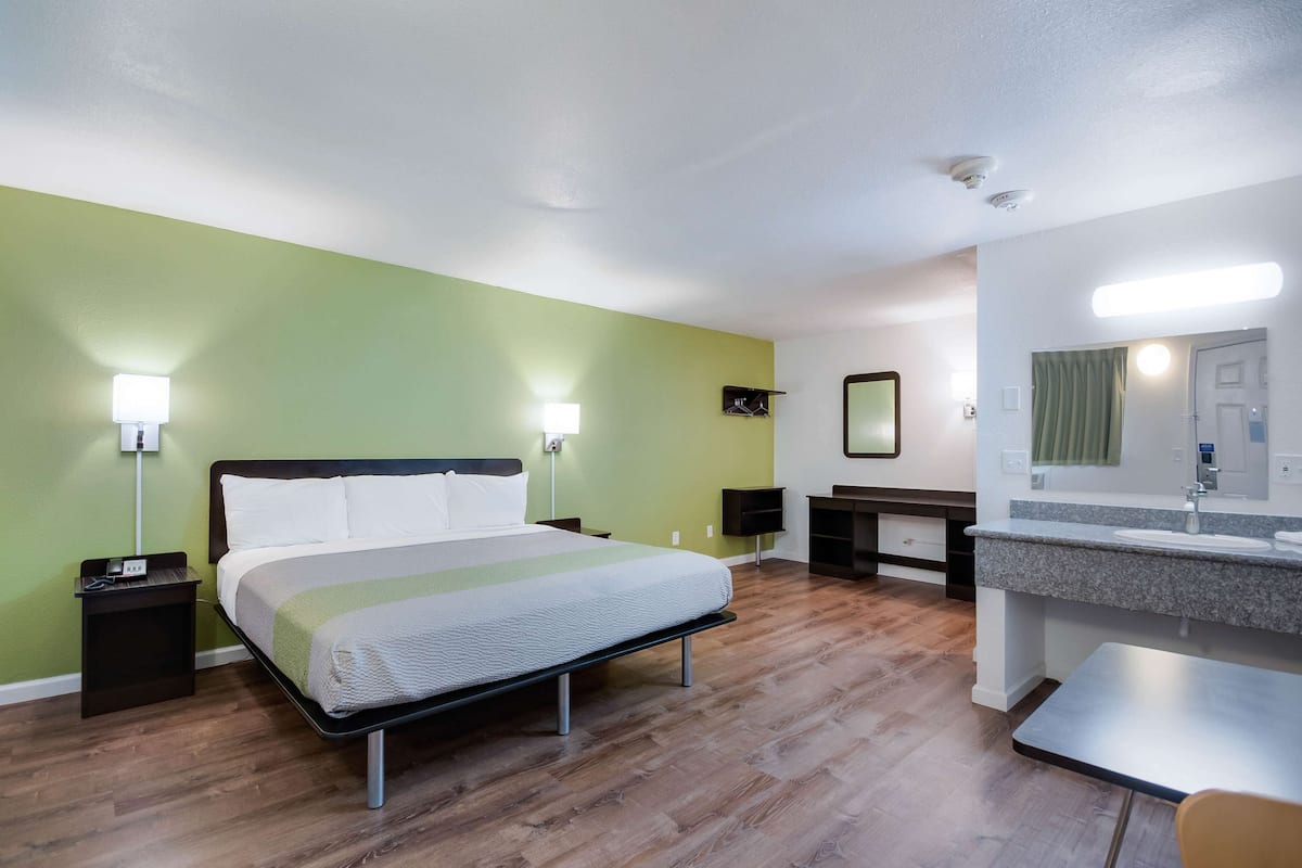 A hotel room with a bed, bedside tables, a desk, a chair, a mirror, and a bathroom with a sink and mirror.
