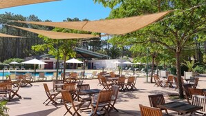 Een buitenzwembad met houten stoelen en tafels, beschaduwd door grote parasols, en een gebouw op de achtergrond.