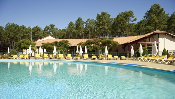 A swimming pool with yellow lounge chairs and white umbrellas, surrounded by trees and a building with a tiled roof. - Village Vacances Le Lac Marin Soustons (Soustons)