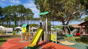 A playground with slides and climbing structures, surrounded by trees and a building in the background. - Village Vacances Le Lac Marin Soustons (Soustons)