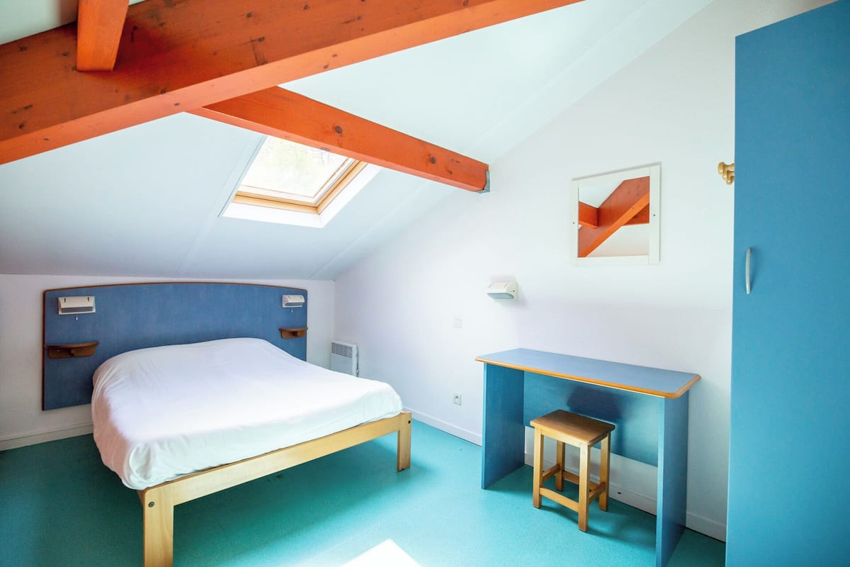 Een slaapkamer met een bed, een bureau, een krukje, een kledingkast en een dakraam.