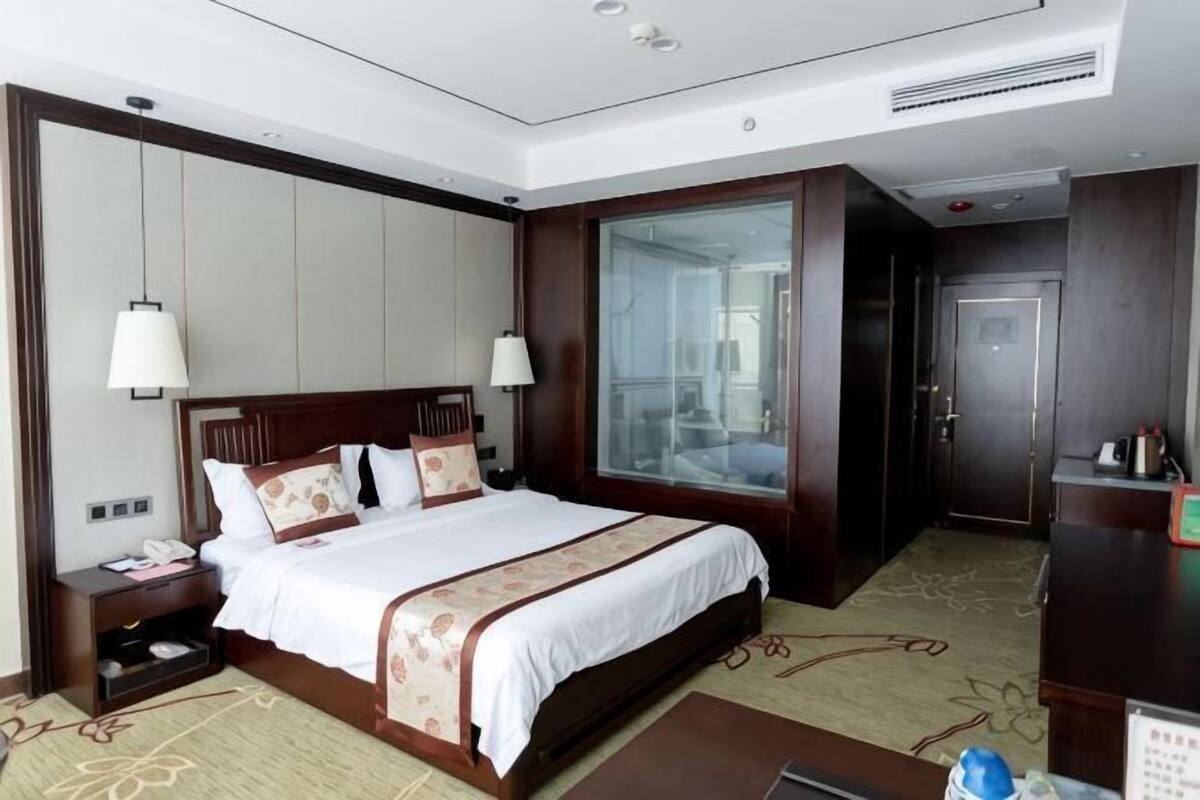A hotel room with a large bed, bedside lamps, a nightstand, a desk, and a view of the city through a large window.