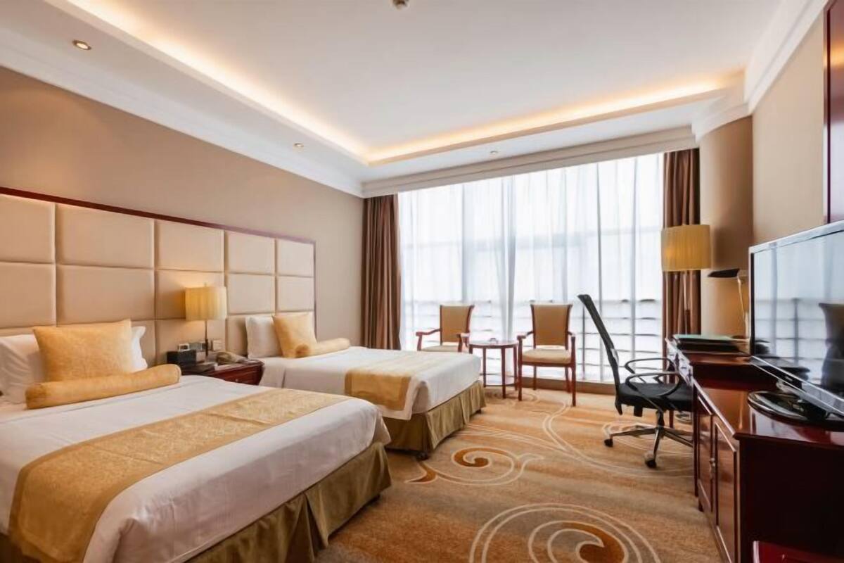 A hotel room with two beds, a desk, a TV, and a large window.