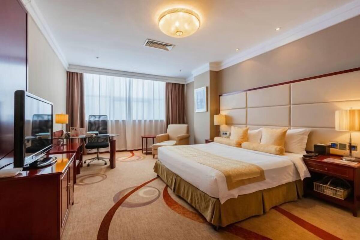 A hotel room with a large bed, a desk with a TV, a chair, and a window with curtains.