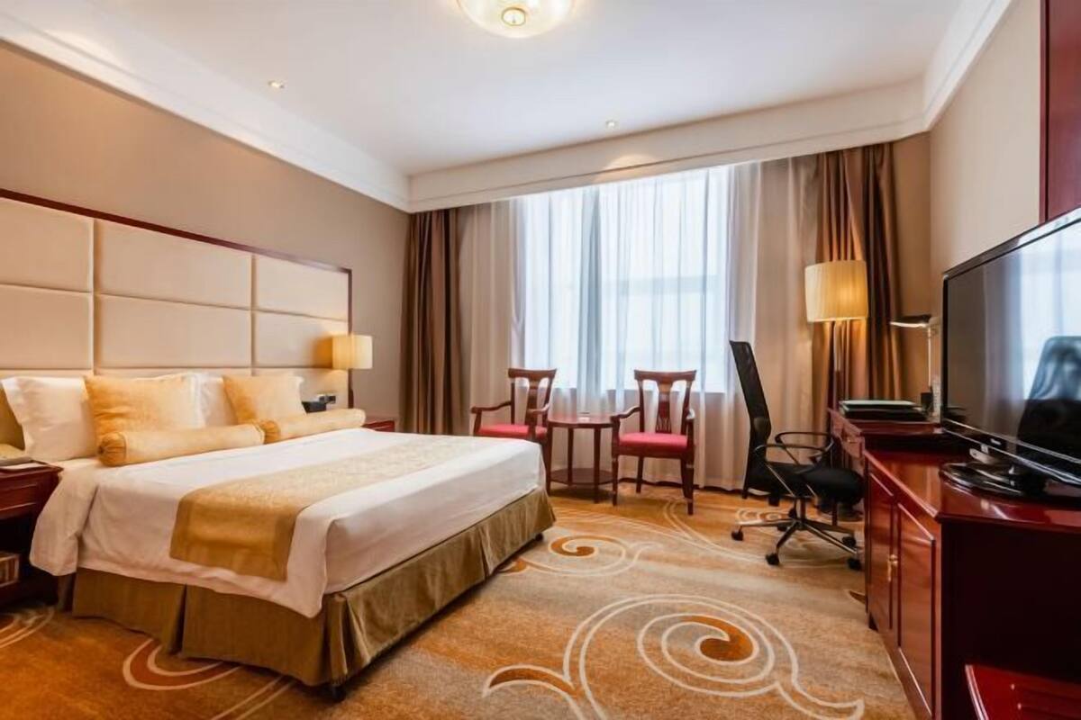 A hotel room with a large bed, a desk, a chair, a TV, and a window with curtains.