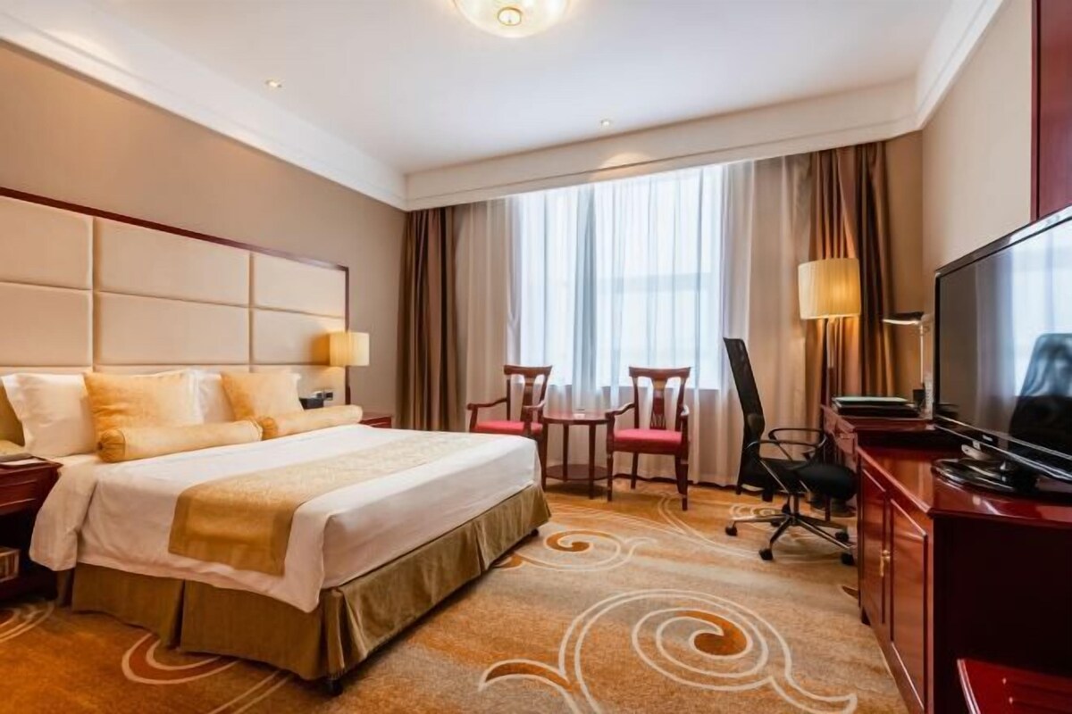 A hotel room with a large bed, a desk, a chair, a TV, and a window with curtains.