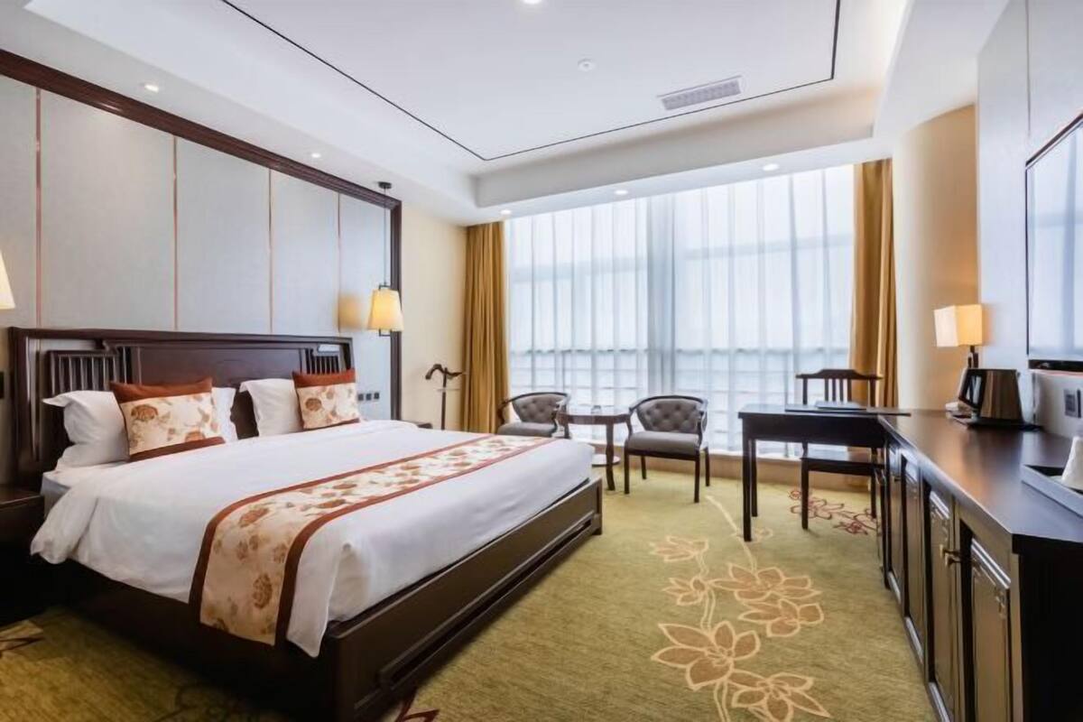 A hotel room with a large bed, a desk, two chairs, and a view of the city through large windows.