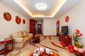 Quarto de hotel com sofá, televisão, flores decorativas e arte tradicional nas paredes.
