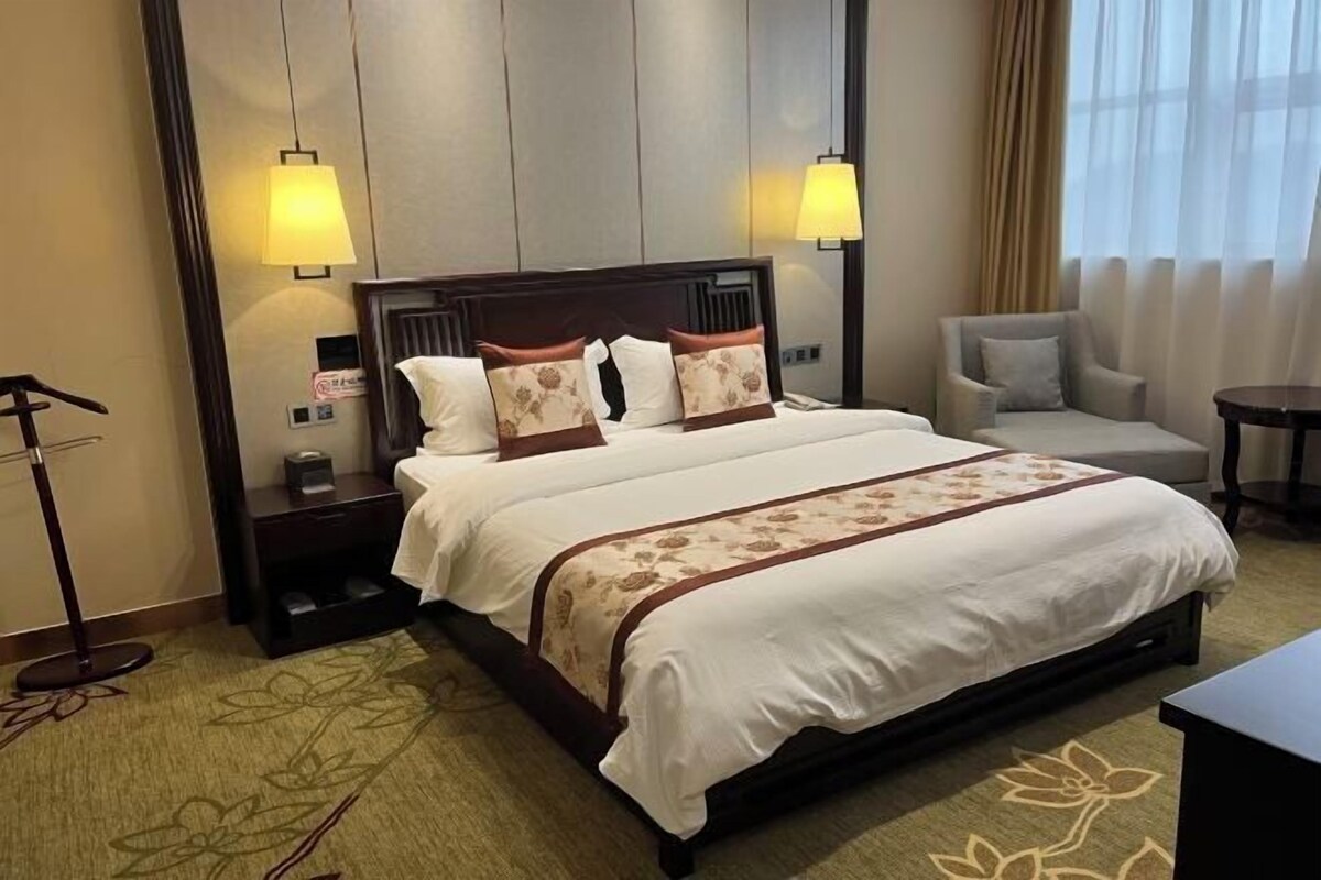 A hotel room with a large bed, two bedside lamps, a chair, a desk, and a carpet with a floral pattern.
