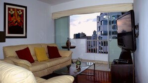 A living room with a beige sofa, red and yellow cushions, a glass coffee table, a TV on a stand, and a city view through a window. - The Comfortable Flat Miraflores (Lima)