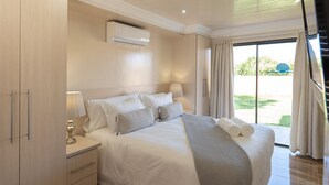 Una habitación de hotel con una cama grande, una mesita de noche con una lámpara, un equipo de aire acondicionado y vistas al exterior a través de un ventanal corredero.