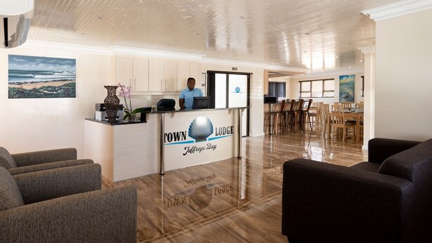 Zona de recepción con un cartel que dice "Town Lodge Jebreys Bay", una recepcionista y un comedor con sillas y mesas.