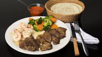 Un plato de comida con camarones, bistec, brócoli y zanahorias, una cesta de tortillas y un vaso de salsa sobre una mesa negra.