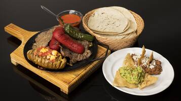 Una tabla de cortar de madera con una sartén negra que contiene un chile relleno, una cesta de tortillas y un plato con guacamole y frijoles.