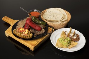 Una tabla de cortar de madera con una sartén negra que contiene un chile relleno, una cesta de tortillas y un plato con guacamole y frijoles.