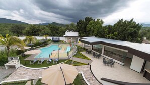 Un área de piscina moderna al aire libre con un salón con paredes de cristal, una piscina con tobogán y vistas de la vegetación y montañas circundantes.