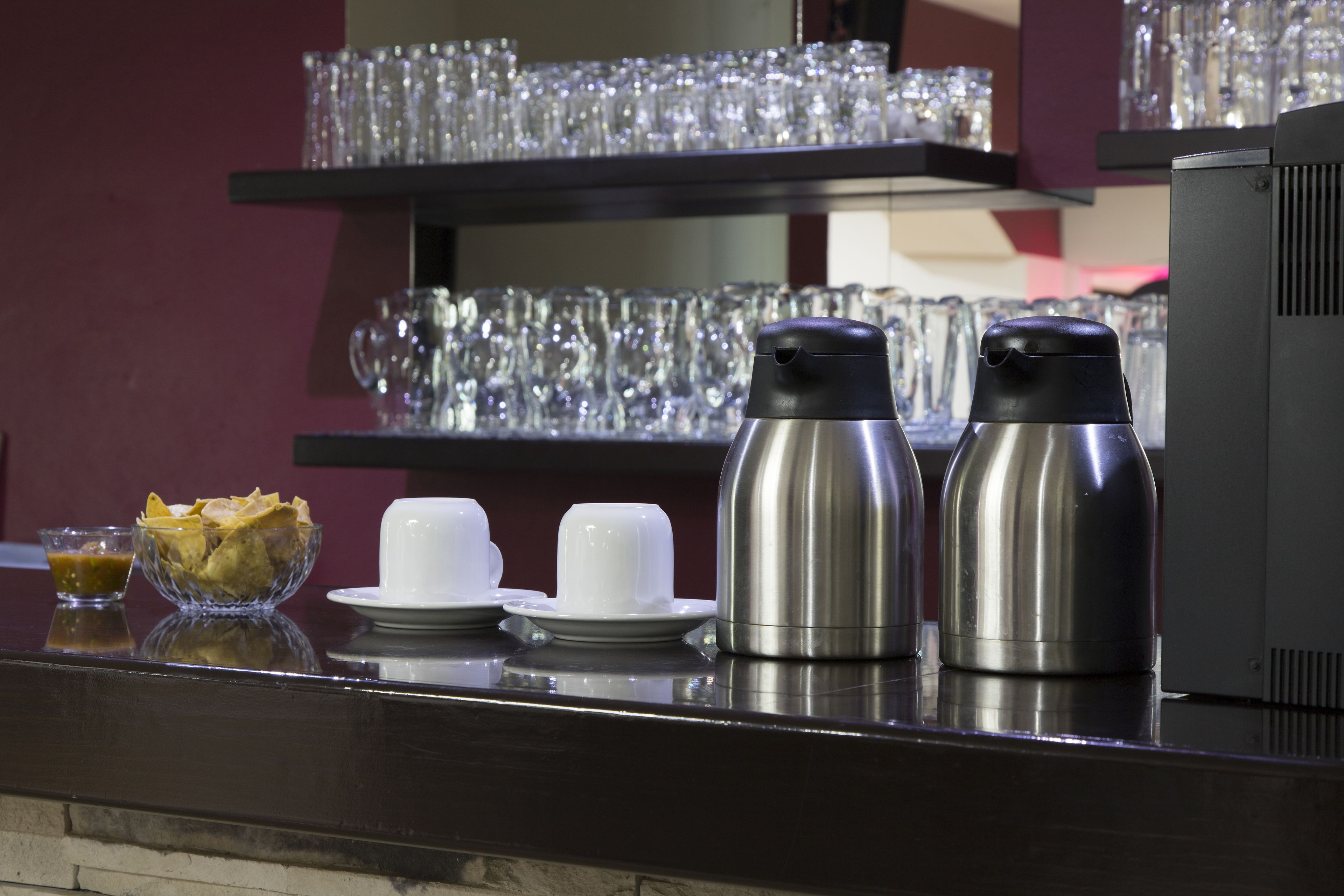 Una zona de bar con una máquina de café, un bol de patatas fritas y varias tazas y platillos blancos.
