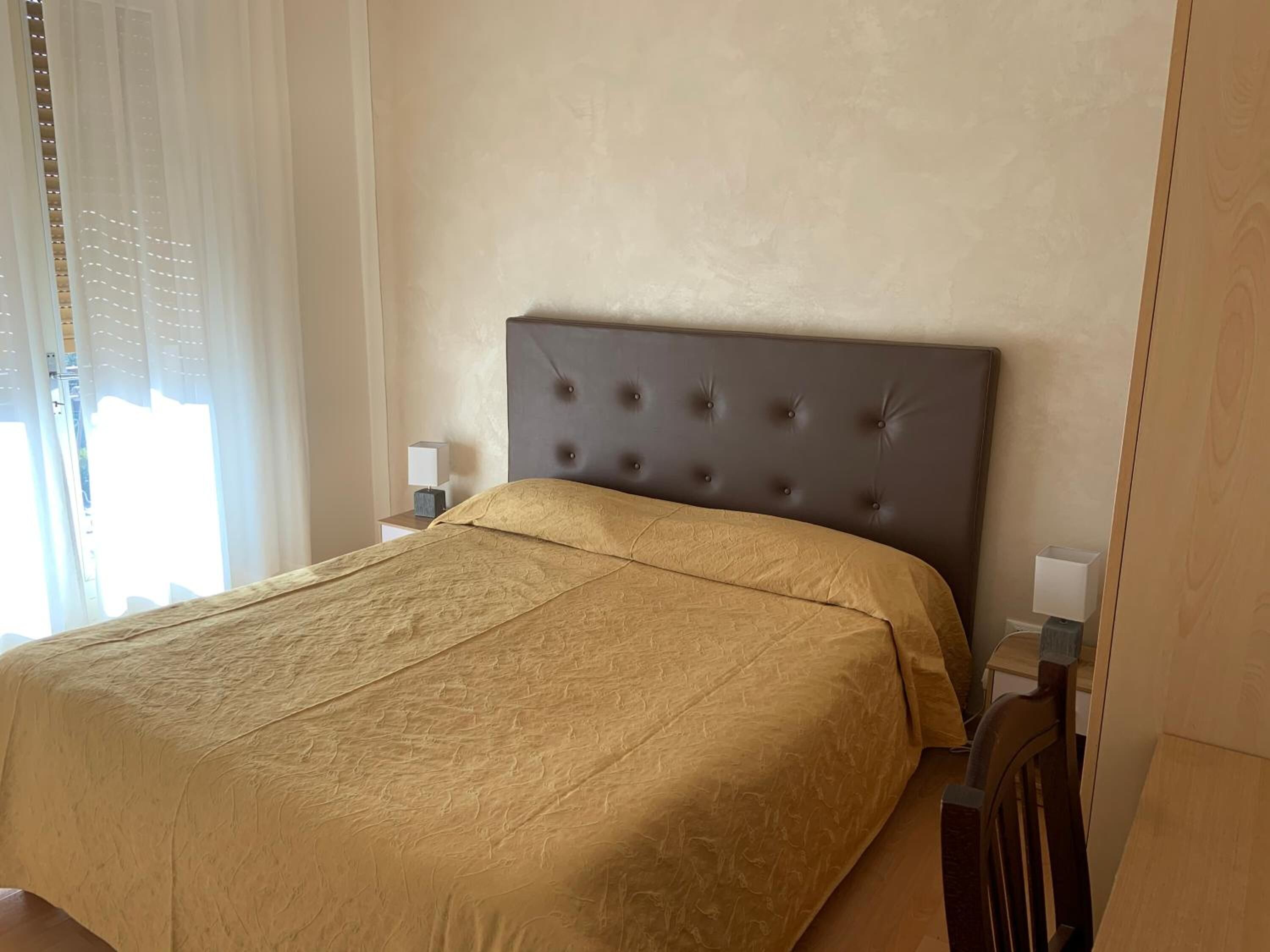 A neatly made bed with a tufted headboard, a bedside table with a lamp, and a window with sheer curtains.