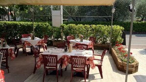 Un espacio de comedor al aire libre con mesas y sillas, cubierto por un dosel, rodeado de follaje.
