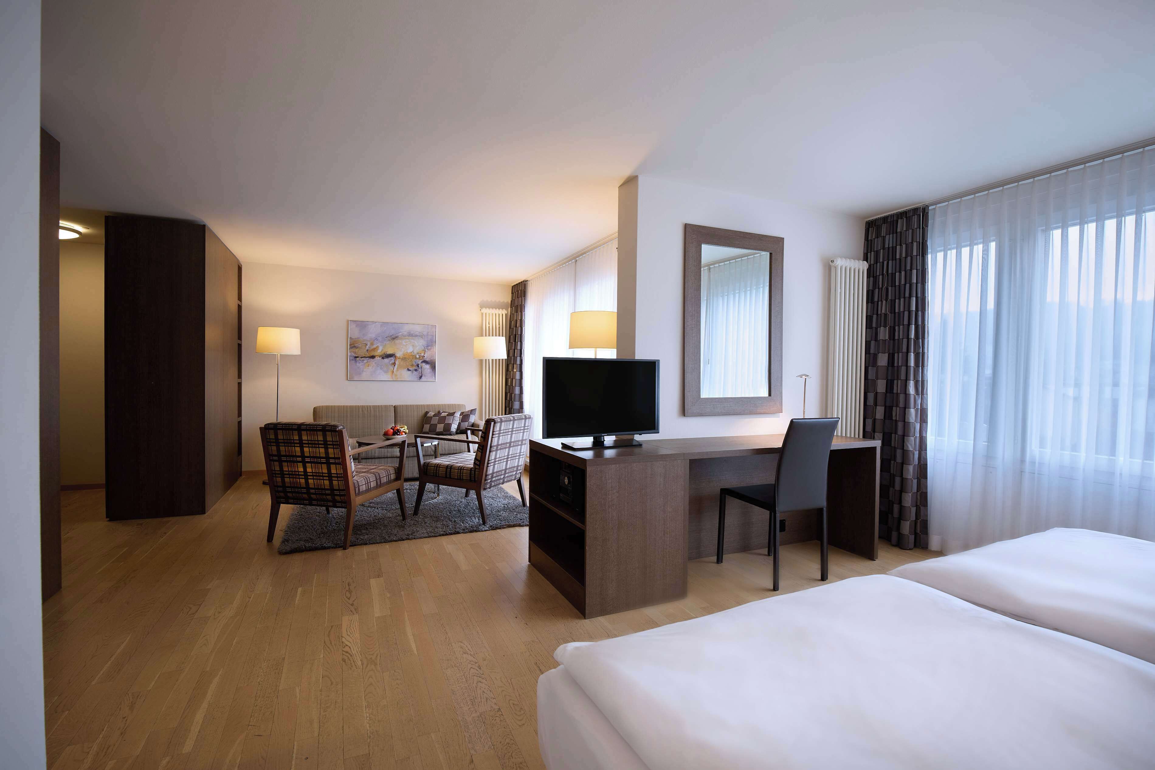 Habitación de hotel con una cama grande, un escritorio con televisor, una silla, un sofá y vistas al exterior por la ventana.