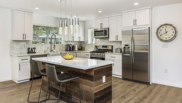 Una cocina moderna con armarios blancos, electrodomésticos de acero inoxidable, una isla de madera con taburetes altos y un reloj en la pared.