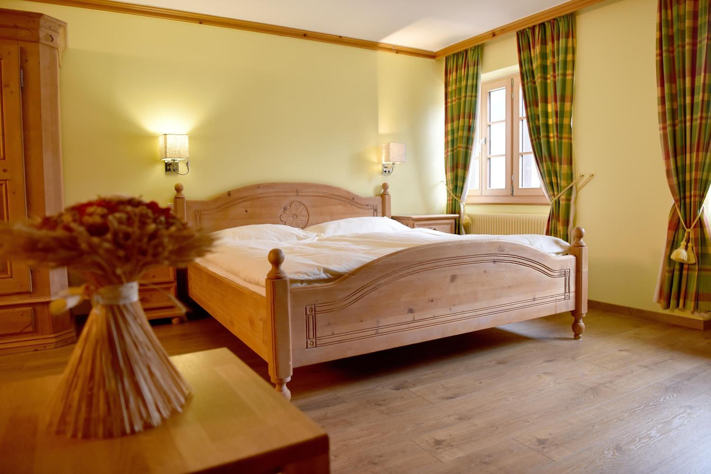 Un dormitorio con una cama de madera, dos lámparas de noche, un ventanal con cortinas y un jarrón con flores secas sobre una mesa de madera.