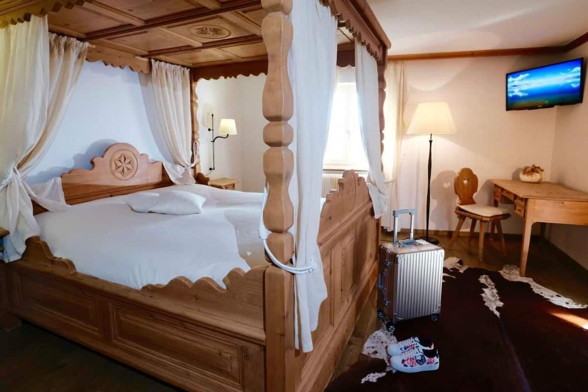 A four-poster canopy bed with a wooden headboard, a bedside table with a lamp, a desk with a chair, and a television on the wall.