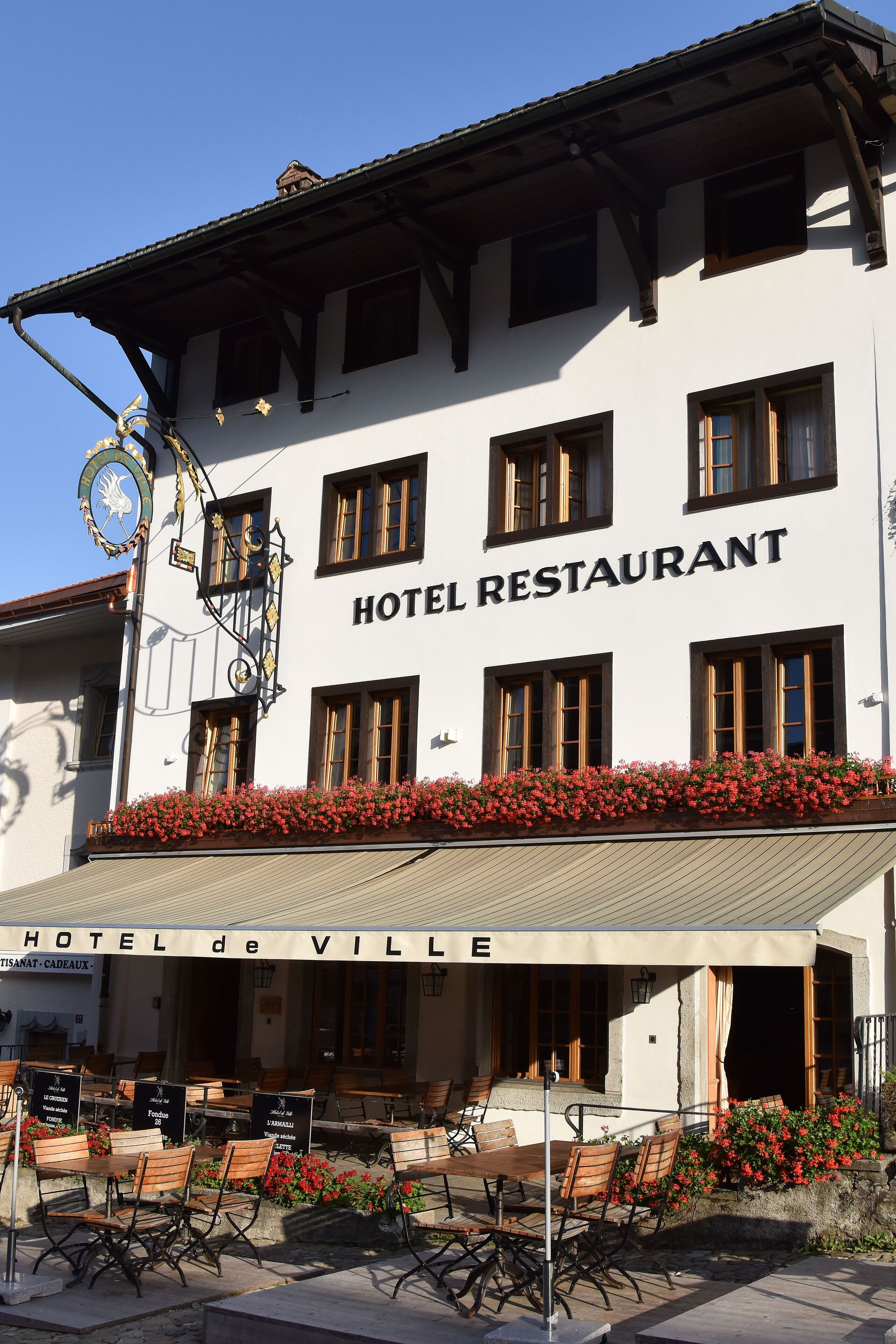 A hotel with a sign reading "HOTEL RESTAURANT" and "HOTEL de VILLE". It has outdoor seating with tables and chairs, and a flower bed with red flowers.