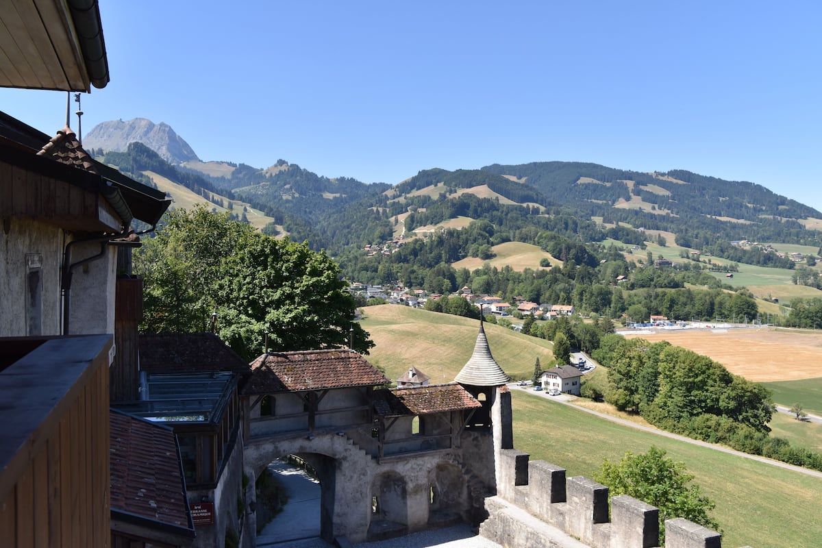 A historic building with a stone facade and a tower, overlooking a valley with rolling hills and a mountain in the background.