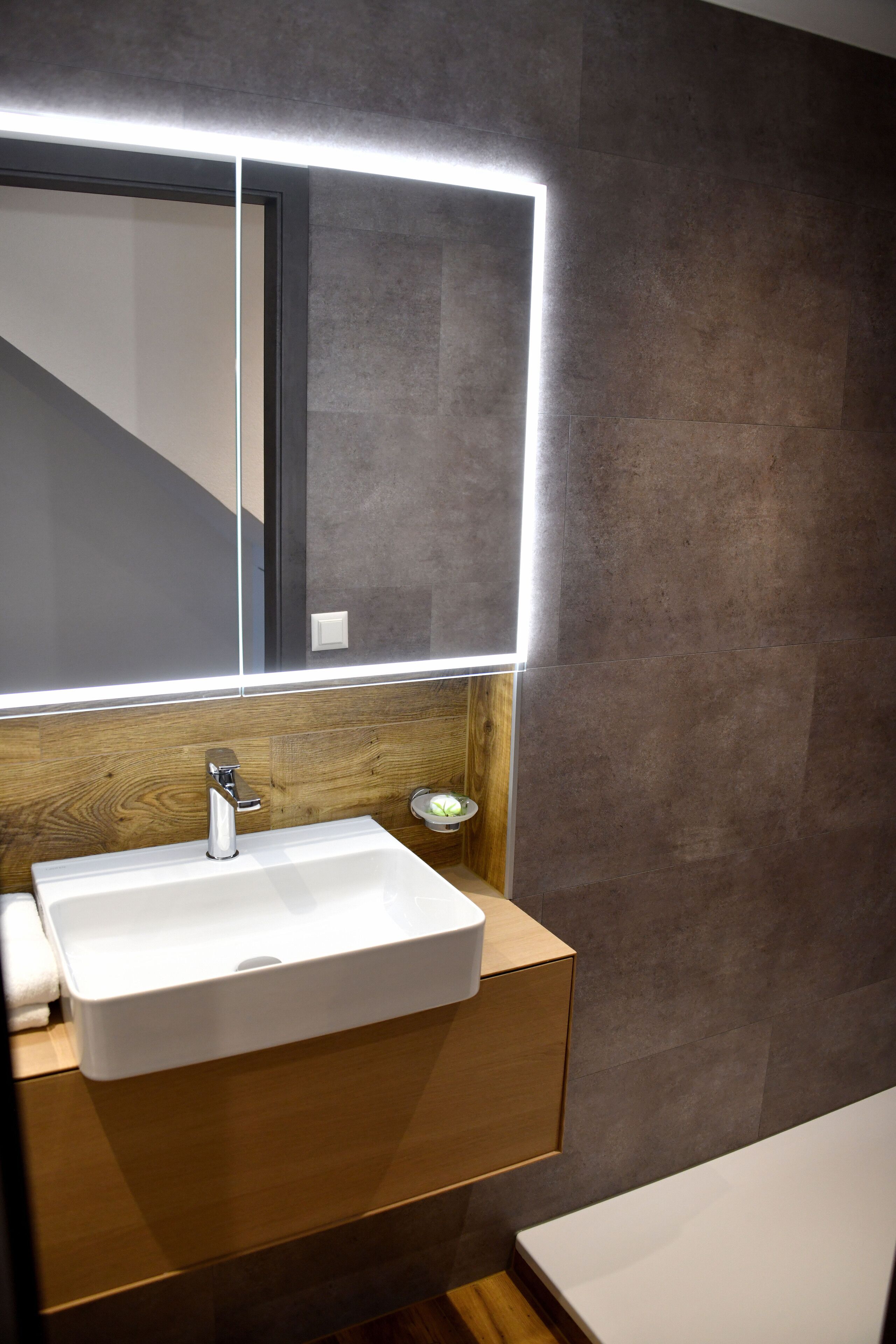 Une salle de bain moderne avec un lavabo blanc, un meuble-lavabo en bois et un grand miroir doté d’un éclairage intégré.