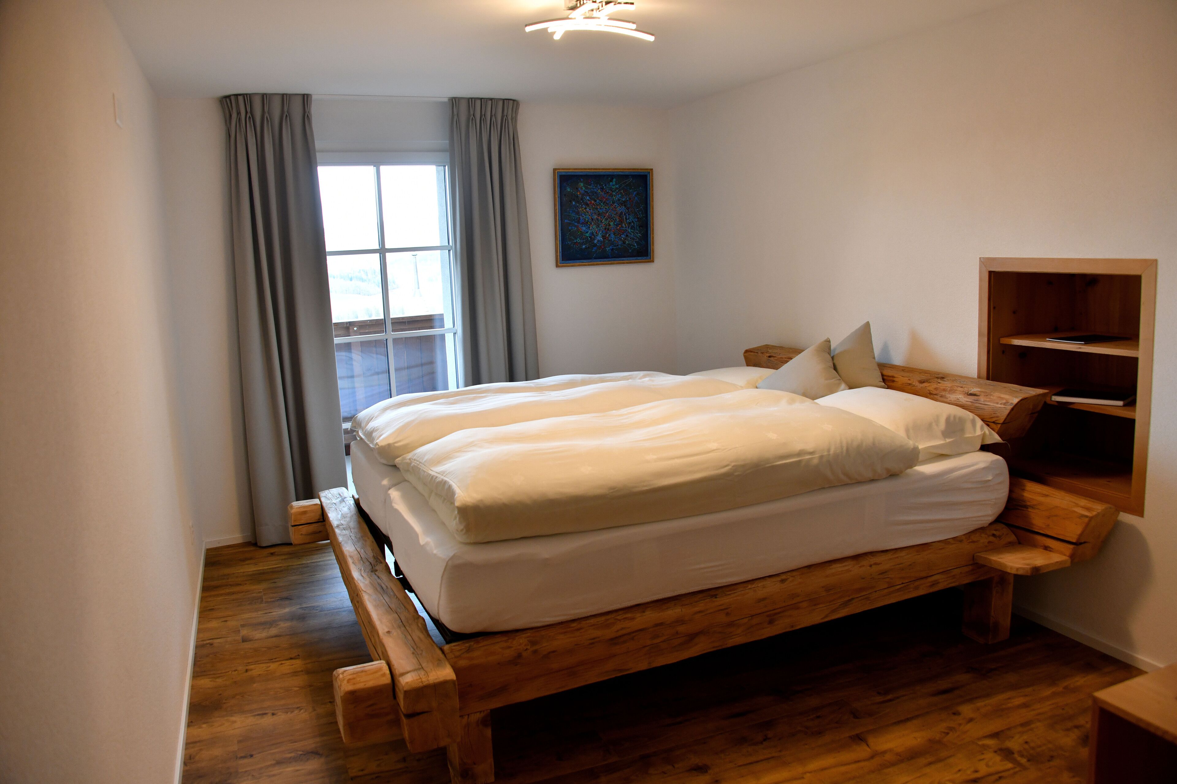Un dormitorio con una cama de madera, una ventana con cortinas, un cuadro en la pared y una estantería de madera.