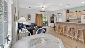 Un salon comprenant un canapé, des fauteuils, une table à manger et un coin cuisine avec des tabourets et des placards.