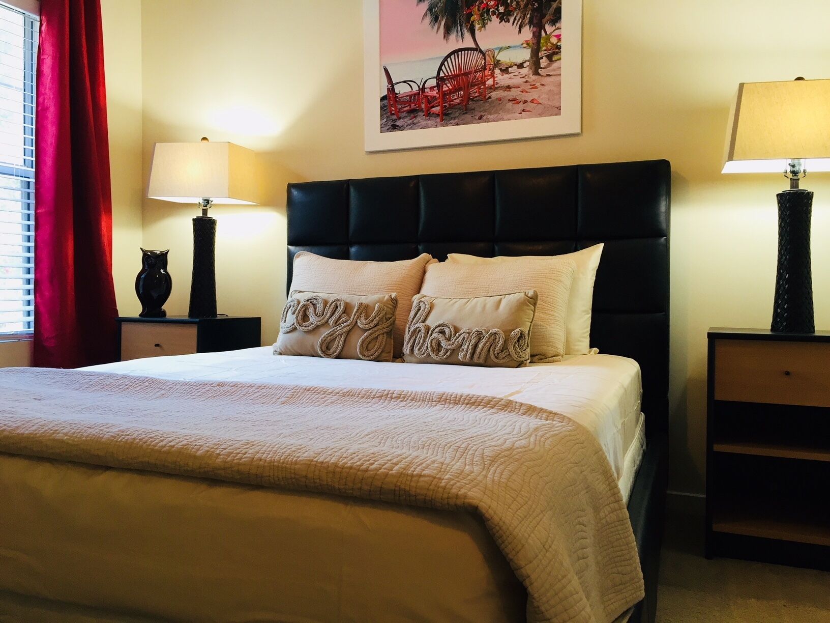 A neatly made bed with a headboard, two bedside tables with lamps, a framed picture on the wall, and a window with red curtains.