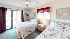 Quarto com duas camas, um ventilador de teto, janela com cortinas, mesa de cabeceira com abajur e cômoda.
