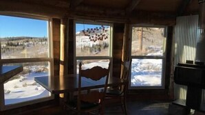 Interior de una cabaña de madera con mesa y sillas, ventanas amplias con vistas nevadas y chimenea.
