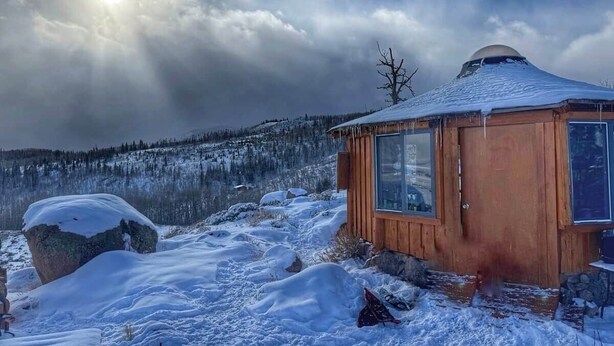 Una cabaña de madera con techo nevado, rodeada por un paisaje invernal y una cadena montañosa cubierta de bosques.