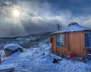 Eine Holzhütte mit schneebedecktem Dach, umgeben von verschneiter Landschaft und einem bewaldeten Gebirgszug.