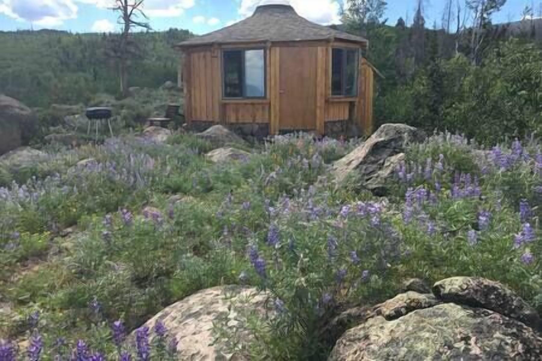 A wooden cabin with a pitched roof, surrounded by a natural landscape with rocks and purple flowers.