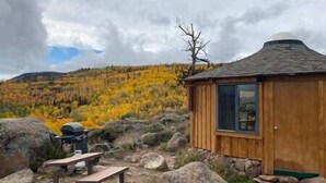 Una cabaña de madera con un solo ventanal y techo de paja, rodeada de árboles y rocas de tonos otoñales.