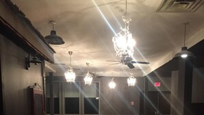 A spacious room with wooden flooring, multiple chandeliers, ceiling fans, and a visible exit sign. - Hamilton Hotel (Williams Lake)